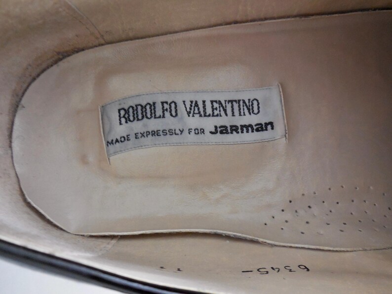 Puede incluir: Vista interior de un zapato negro con una plantilla color crema. Una etiqueta rectangular dice "RODOLFO VALENTINO MADE EXPRESSLY FOR JARMAN" en texto negro. La plantilla del zapato tiene peque&ntilde;os agujeros de ventilaci&oacute;n.