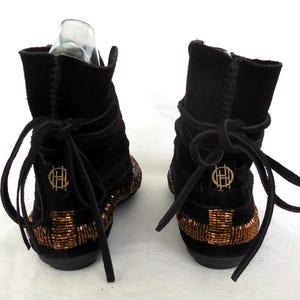 K&ouml;nnte beinhalten: Paar schwarze Wildleder-Ankle Boots mit kupferfarbenen Perlenarbeiten entlang der Sohlen. Die Stiefel haben ein Wickeldesign mit Schn&uuml;rverschl&uuml;ssen und einem goldenen Emblem auf der R&uuml;ckseite. Die Stiefel werden vor wei&szlig;em Hintergrund pr&auml;sentiert.