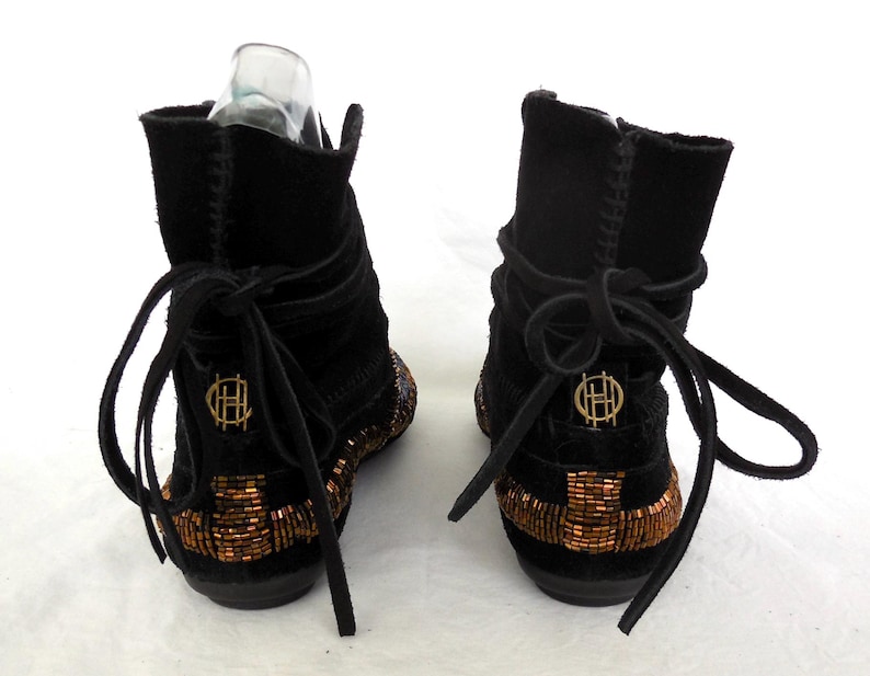 K&ouml;nnte beinhalten: Paar schwarze Wildleder-Ankle Boots mit kupferfarbenen Perlenarbeiten entlang der Sohlen. Die Stiefel haben ein Wickeldesign mit Schn&uuml;rverschl&uuml;ssen und einem goldenen Emblem auf der R&uuml;ckseite. Die Stiefel werden vor wei&szlig;em Hintergrund pr&auml;sentiert.