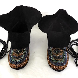 K&ouml;nnte beinhalten: Ein Paar schwarze Wildleder-Mokassin-Stiefel mit Perlenmustern in Blau, Gr&uuml;n und Orange. Die Stiefel haben einen umgeschlagenen Schaft und lange schwarze Schn&uuml;rsenkel.