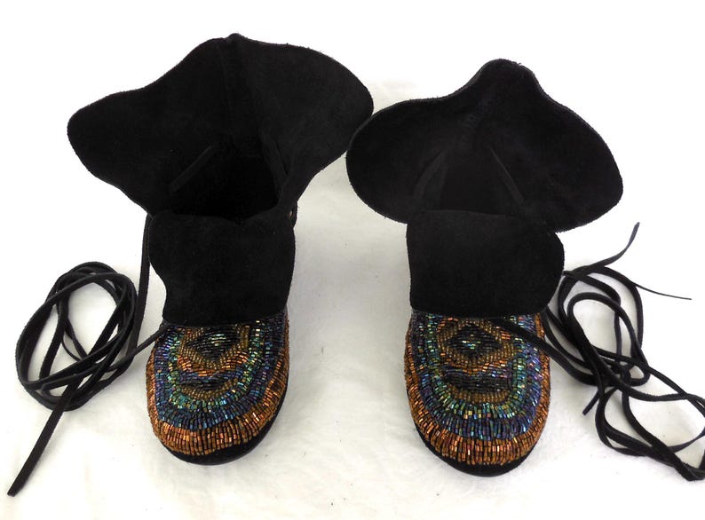 K&ouml;nnte beinhalten: Ein Paar schwarze Wildleder-Mokassin-Stiefel mit Perlenmustern in Blau, Gr&uuml;n und Orange. Die Stiefel haben einen umgeschlagenen Schaft und lange schwarze Schn&uuml;rsenkel.