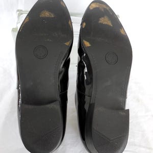 Puede incluir: Un par de zapatos de charol negro, vistos desde las suelas. Las suelas son negras con algo de desgaste, y un logotipo circular es visible en cada suela. Los tacones tienen un dise&ntilde;o texturizado.