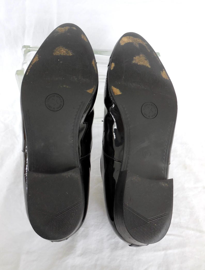 Puede incluir: Un par de zapatos de charol negro, vistos desde las suelas. Las suelas son negras con algo de desgaste, y un logotipo circular es visible en cada suela. Los tacones tienen un dise&ntilde;o texturizado.