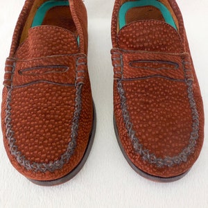 May include: A pair of brown loafers featuring a textured, pebbled surface and a classic penny slot strap across the vamp. The shoes have a moccasin-style stitched toe seam and a bright turquoise interior lining visible at the heel and collar area.