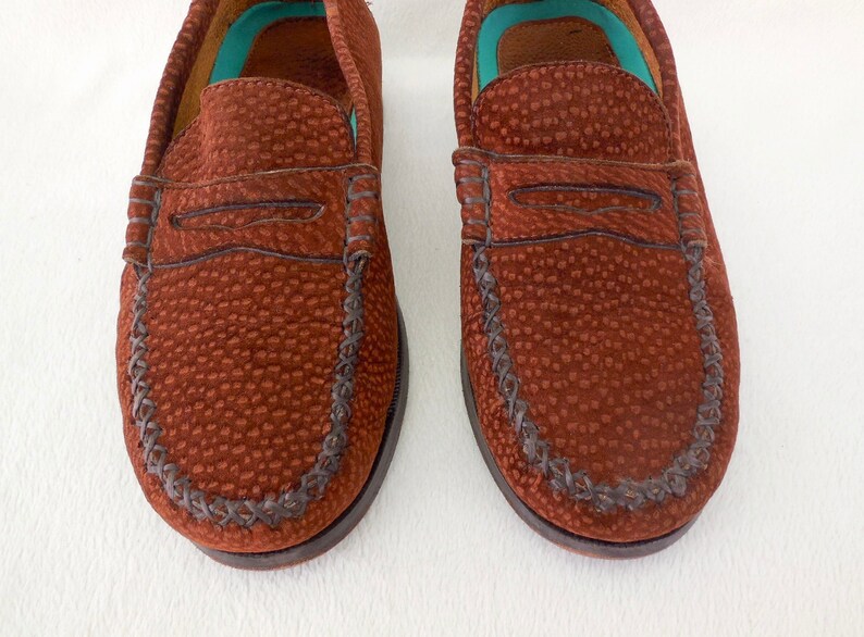 May include: A pair of brown loafers featuring a textured, pebbled surface and a classic penny slot strap across the vamp. The shoes have a moccasin-style stitched toe seam and a bright turquoise interior lining visible at the heel and collar area.