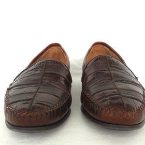 K&ouml;nnte beinhalten: Ein Paar braune Lederschuhe mit Kroko-Pr&auml;gung. Die Schuhe haben einen gen&auml;hten Rand und eine spitze Zehenpartie.