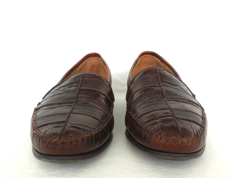 K&ouml;nnte beinhalten: Ein Paar braune Lederschuhe mit Kroko-Pr&auml;gung. Die Schuhe haben einen gen&auml;hten Rand und eine spitze Zehenpartie.