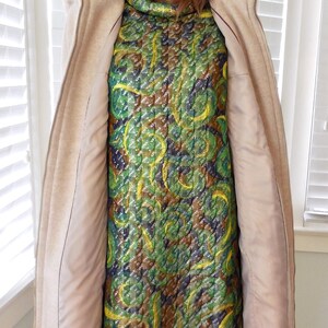 May include: A mannequin displays a vintage dress and coat. The dress features a quilted pattern in green, yellow, and brown. A beige zip-up coat with a high collar is worn over the dress. The coat's interior is a light pink.