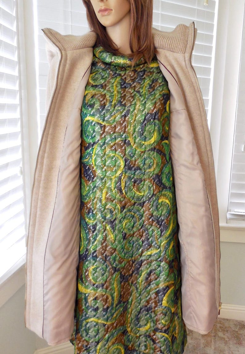 May include: A mannequin displays a vintage dress and coat. The dress features a quilted pattern in green, yellow, and brown. A beige zip-up coat with a high collar is worn over the dress. The coat's interior is a light pink.