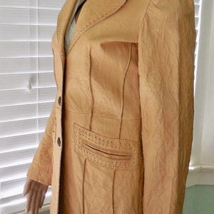 May include: A tan leather jacket with a long, fitted silhouette. The jacket has a notched collar, a single-breasted closure, and two front pockets. The jacket is decorated with stitching details.
