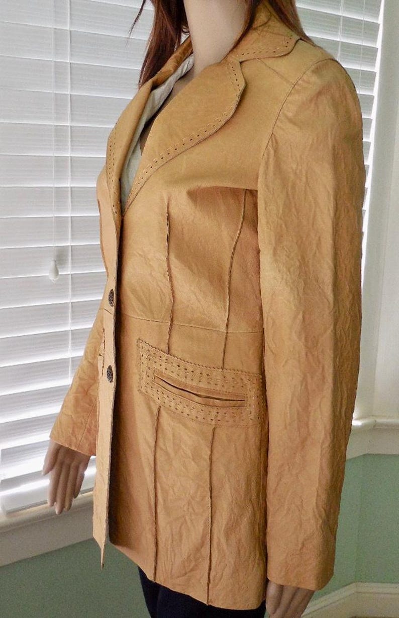 May include: A tan leather jacket with a long, fitted silhouette. The jacket has a notched collar, a single-breasted closure, and two front pockets. The jacket is decorated with stitching details.