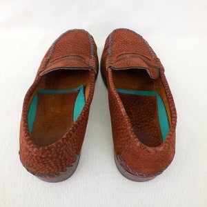 May include: A pair of brown loafers with a textured, pebbled leather exterior and a distinctive stitched moccasin-style toe seam. The interior features a smooth brown leather lining with a contrasting bright turquoise insole insert.