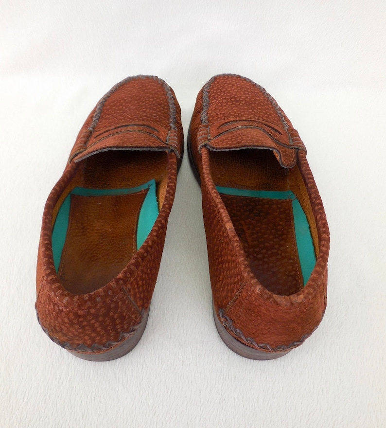 May include: A pair of brown loafers with a textured, pebbled leather exterior and a distinctive stitched moccasin-style toe seam. The interior features a smooth brown leather lining with a contrasting bright turquoise insole insert.