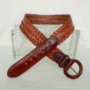 May include: A brown leather belt with a braided design and a silver buckle. The belt is made of woven leather and has a smooth, textured finish.