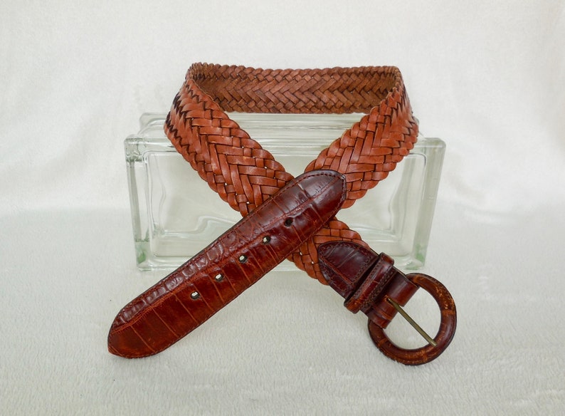 May include: A brown leather belt with a braided design and a silver buckle. The belt is made of woven leather and has a smooth, textured finish.