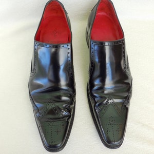 Puede incluir: Un par de zapatos negros de cuero sin cordones con punta puntiaguda y costuras decorativas. Los zapatos tienen un forro de cuero rojo.