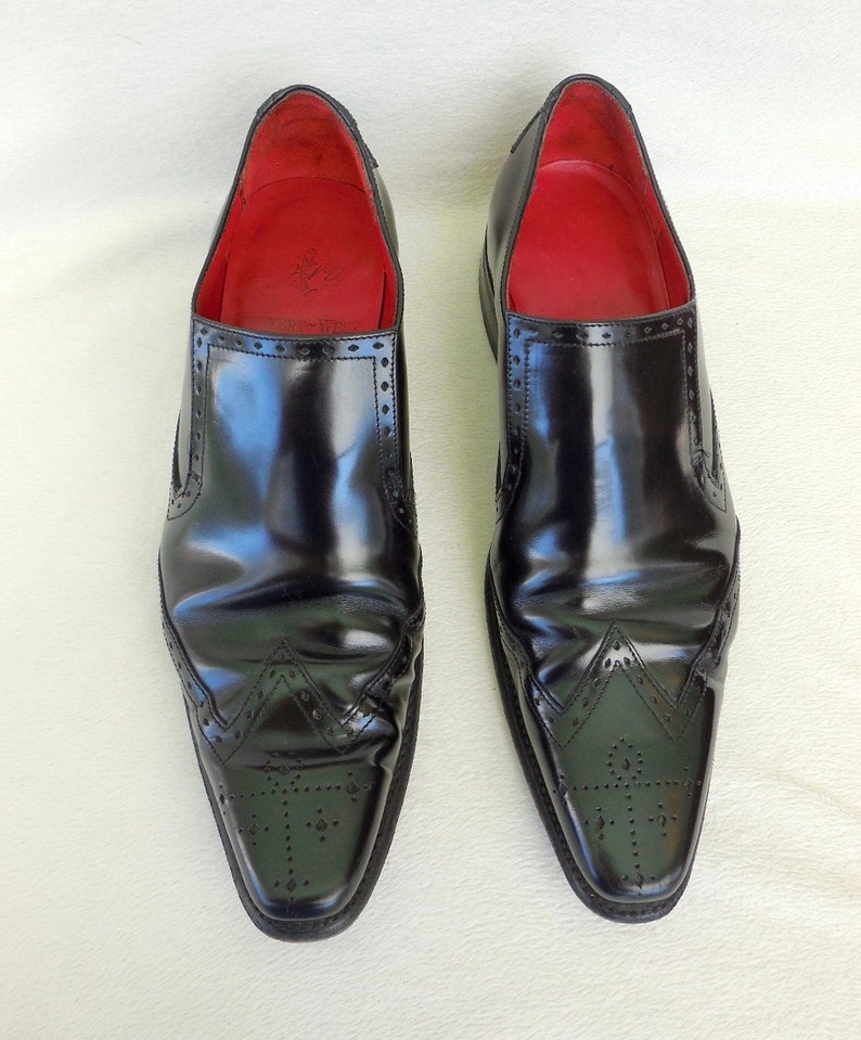 Puede incluir: Un par de zapatos negros de cuero sin cordones con punta puntiaguda y costuras decorativas. Los zapatos tienen un forro de cuero rojo.