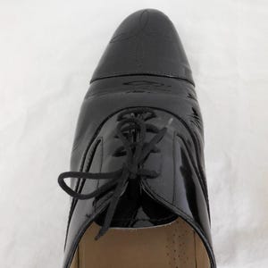 Puede incluir: Primer plano de un zapato de charol negro con dise&ntilde;o de cordones. El zapato tiene un acabado brillante y costuras decorativas en la puntera. Los cordones son negros y el zapato parece sin usar.