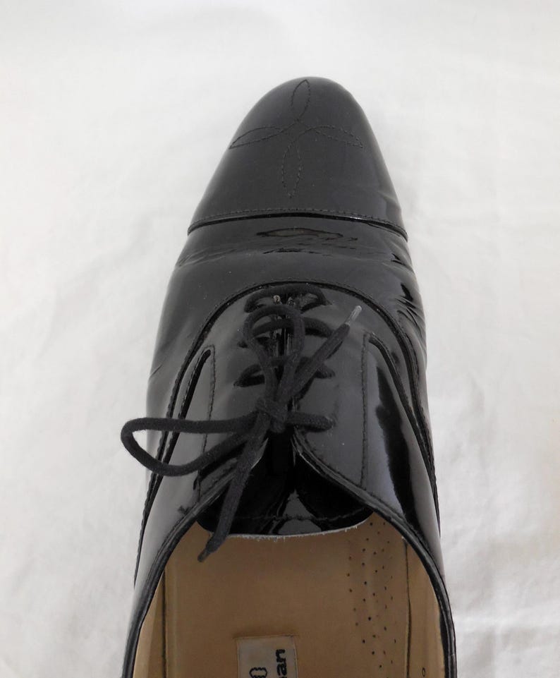 Puede incluir: Primer plano de un zapato de charol negro con dise&ntilde;o de cordones. El zapato tiene un acabado brillante y costuras decorativas en la puntera. Los cordones son negros y el zapato parece sin usar.