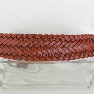 May include: A brown leather belt with a woven design. The belt is lying on a clear glass surface.