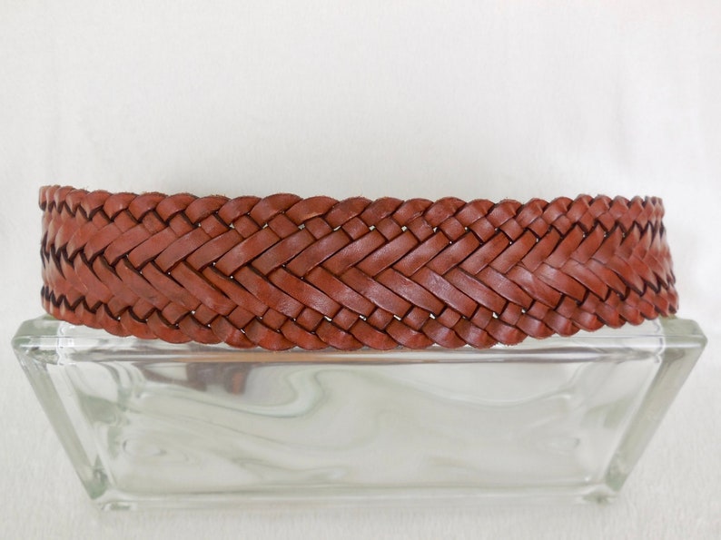 May include: A brown leather belt with a woven design. The belt is lying on a clear glass surface.
