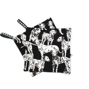 May include: Two black potholders with a Dalmatian dog pattern. The potholders have a black and white Dalmatian print and a loop for hanging. The potholders are made of a quilted fabric.