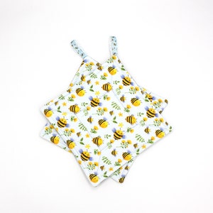 May include: Two white pot holders with a bee and flower pattern. The pot holders have blue and white trim.