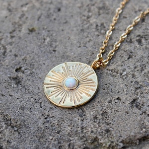 May include: A gold necklace with a round pendant featuring a sunburst design and a white opal stone in the center.