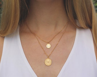 May include: A gold layered necklace with two coin pendants. The larger pendant has a detailed design and the smaller pendant has a simple design.