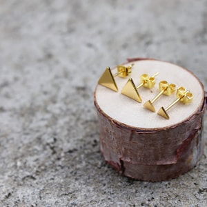 May include: Set of three gold triangle stud earrings displayed on a wooden log. The earrings are different sizes.