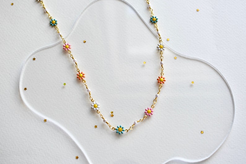 May include: A gold chain necklace with colorful flower charms. The necklace is on a white background with gold glitter.