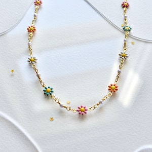 May include: A gold chain necklace with small white, pink, yellow, and blue flower charms. The necklace is on a white background.