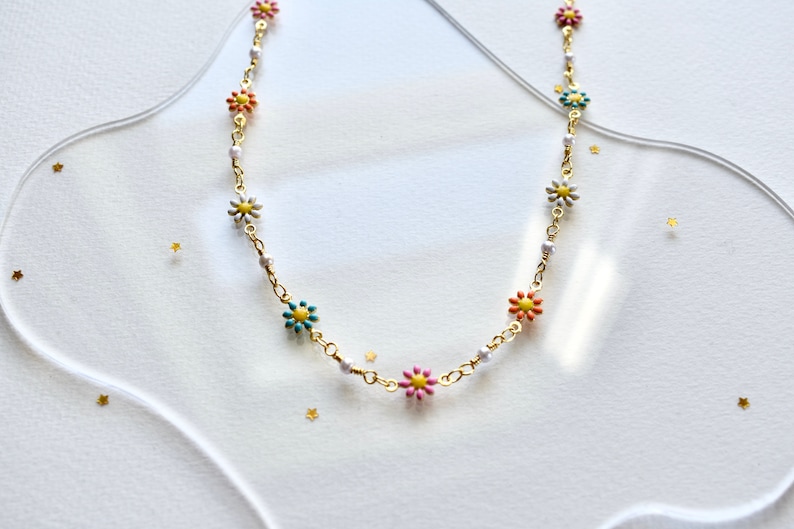May include: A gold chain necklace with small white, pink, yellow, and blue flower charms. The necklace is on a white background.