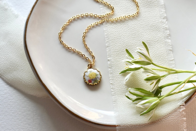 May include: A gold chain necklace with a round pendant featuring a floral design in shades of yellow, red, and green. The pendant is set against a white background and has a scalloped edge.