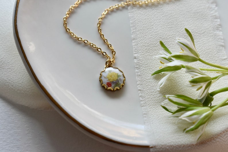 May include: A gold chain necklace with a round pendant featuring a white porcelain cameo with a yellow flower and red berries.