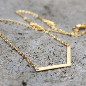 Könnte beinhalten: Eine goldene Kettenkette mit einem einfachen, geometrischen Anhänger in Form eines Chevrons.