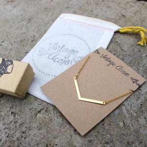 May include: A gold necklace with a chevron pendant on a brown card with the text "Vintage Acorn". The necklace is packaged in a white drawstring bag with the text "Vintage Acorn" in a circle.