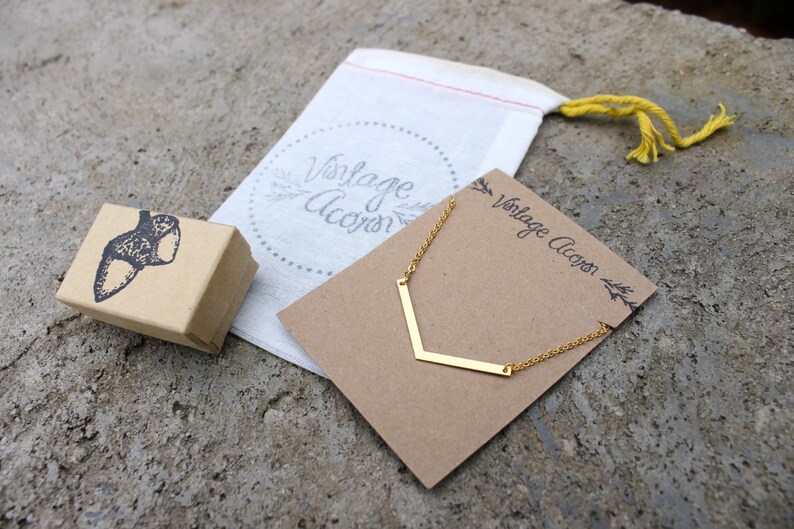 May include: A gold necklace with a chevron pendant on a brown card with the text "Vintage Acorn". The necklace is packaged in a white drawstring bag with the text "Vintage Acorn" in a circle.