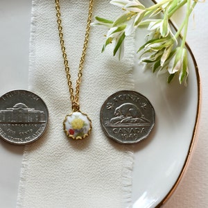 May include: A gold chain necklace with a porcelain pendant featuring a floral design. The pendant is surrounded by a gold trim. The necklace is displayed on a white surface with two coins.
