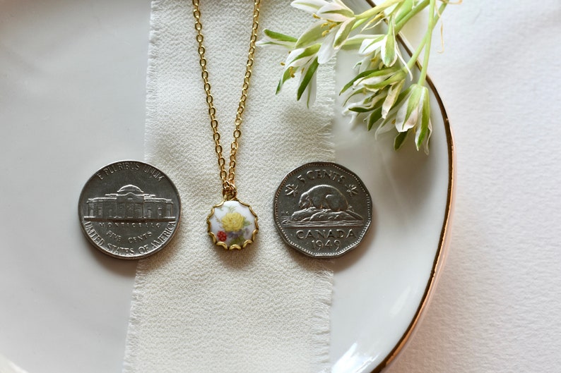 May include: A gold chain necklace with a porcelain pendant featuring a floral design. The pendant is surrounded by a gold trim. The necklace is displayed on a white surface with two coins.