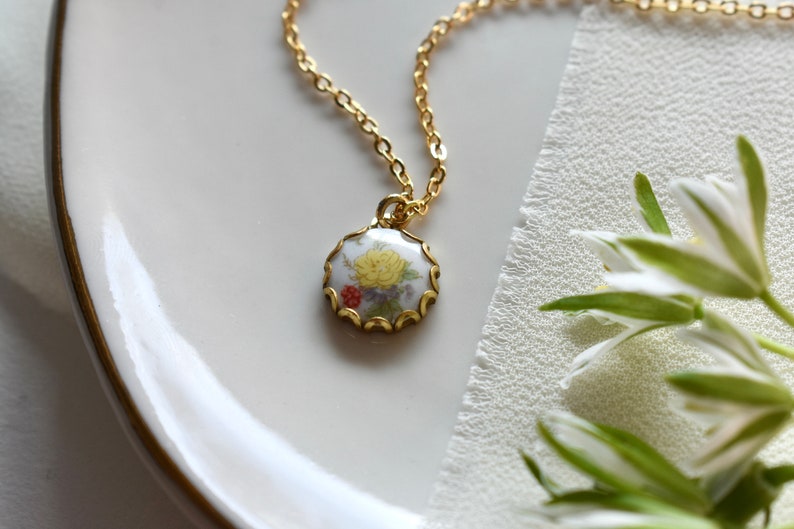 May include: A gold chain necklace with a white porcelain pendant featuring a floral design. The pendant has a scalloped edge and is accented with gold.