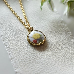 May include: A gold chain necklace with a round pendant featuring a floral design in shades of yellow, red, and green. The pendant is surrounded by a gold filigree frame.
