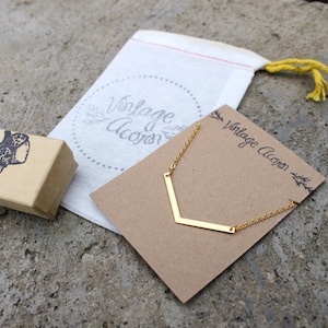 May include: A gold necklace with a chevron pendant on a brown card with the text "Vintage Acorn". The necklace is packaged in a white muslin bag with the text "Vintage Acorn" in a circle.