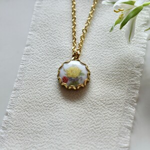 May include: A gold chain necklace with a round pendant featuring a white porcelain cameo with a yellow flower and red and purple accents.