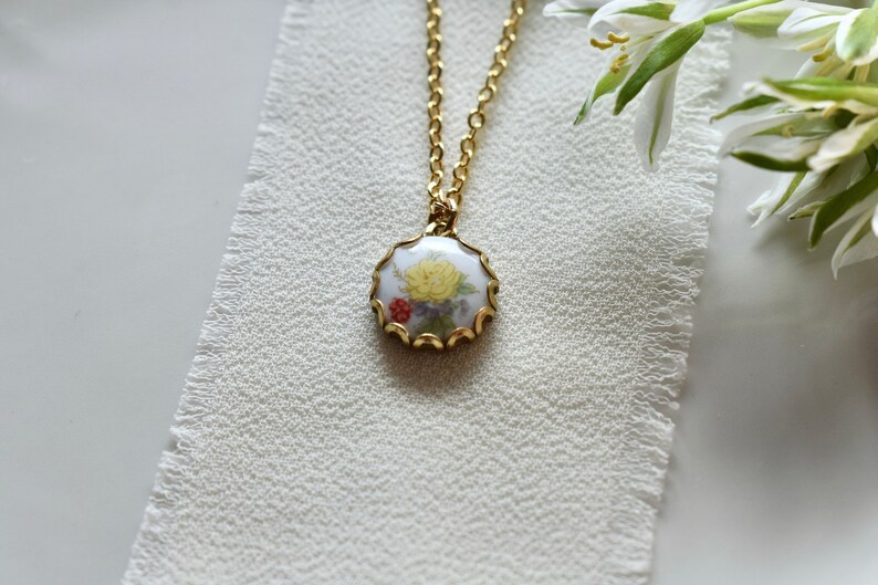 May include: A gold chain necklace with a round pendant featuring a white porcelain cameo with a yellow flower and red and purple accents.