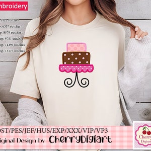 May include: A cream-colored t-shirt featuring a machine embroidery design of a three-tiered cake. The cake has pink and brown layers with polka dots, and sits on a decorative stand. The design is by Cherry Digiart.