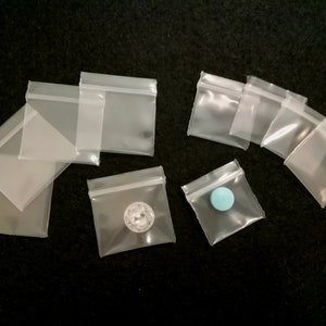 May include: A collection of clear plastic zippered bags of various sizes. Some bags contain small objects, such as a button and a blue pill.
