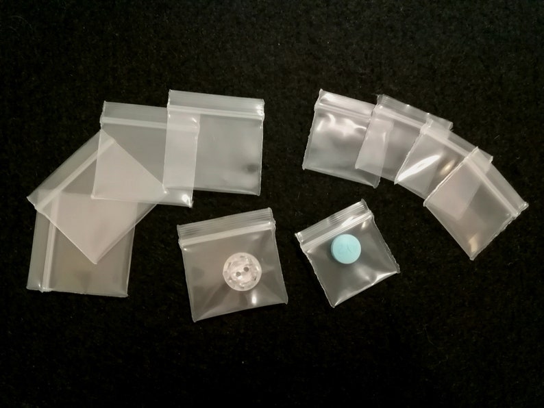 May include: A collection of clear plastic zippered bags of various sizes. Some bags contain small objects, such as a button and a blue pill.