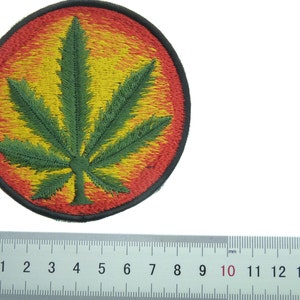 May include: A green marijuana leaf embroidered on a round patch with a red and yellow background. The patch is approximately 5 cm in diameter.