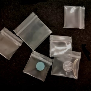 May include: Six clear plastic resealable bags with zipper closures. The bags are various sizes and are lying flat on a black surface. One bag has a small blue circle inside.